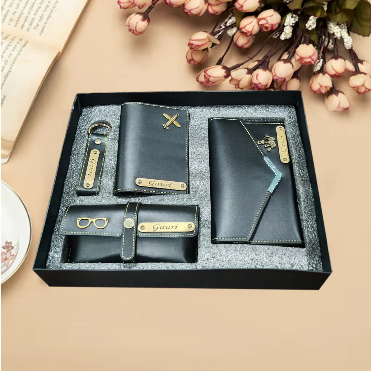 Set of black leather wallets and keychains with 'Gauri' branding in a box on a beige surface with flowers.